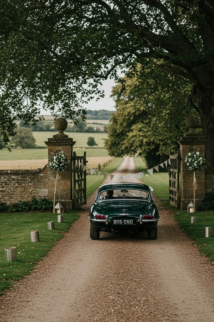 A dark green vintage car driving out through stone gate piers dressed with white flowers, onto a tree-lined drive.