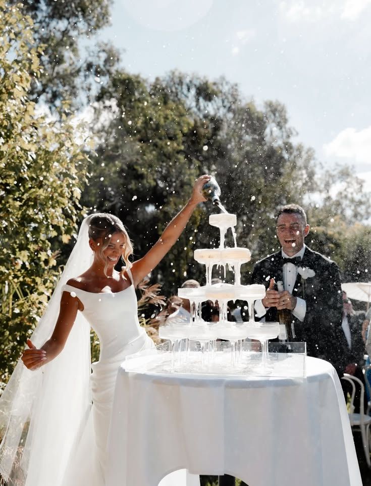 The bride pouring champagne into a tower of coupes in the garden while the groom laughs beside her.