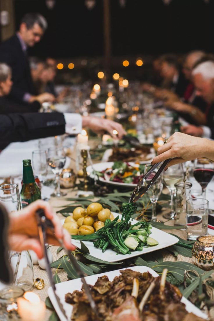 Guests passing platters of food along a long, candlelit table.