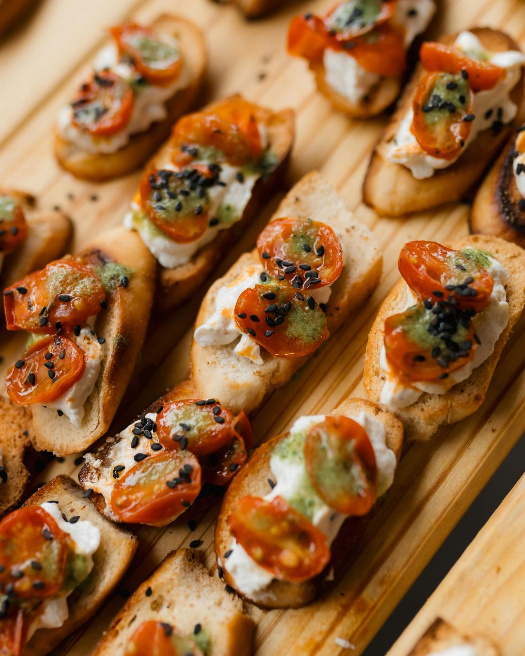 Crostini topped with whipped cheese, roasted tomatoes and black sesame on a wooden board.