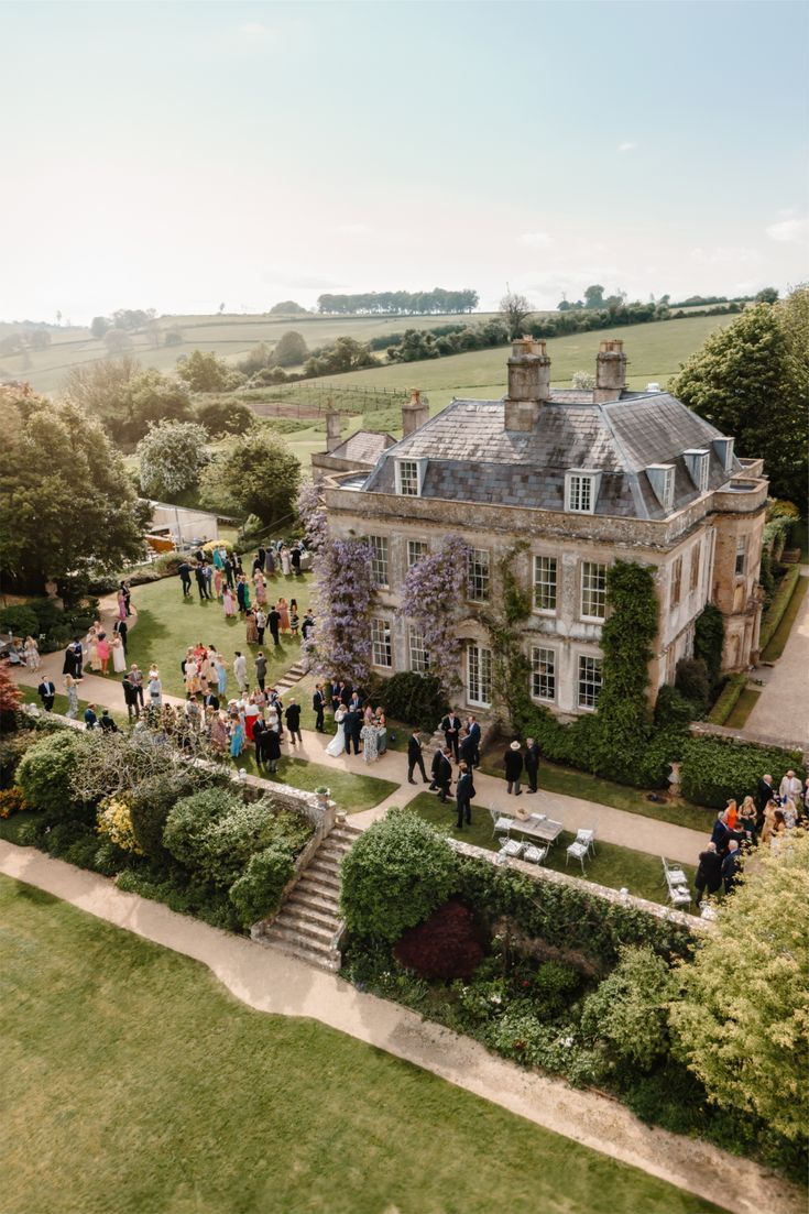 Aerial view of a stone Georgian manor with wisteria on its walls, wedding guests gathered on the lawns, and rolling fields beyond.